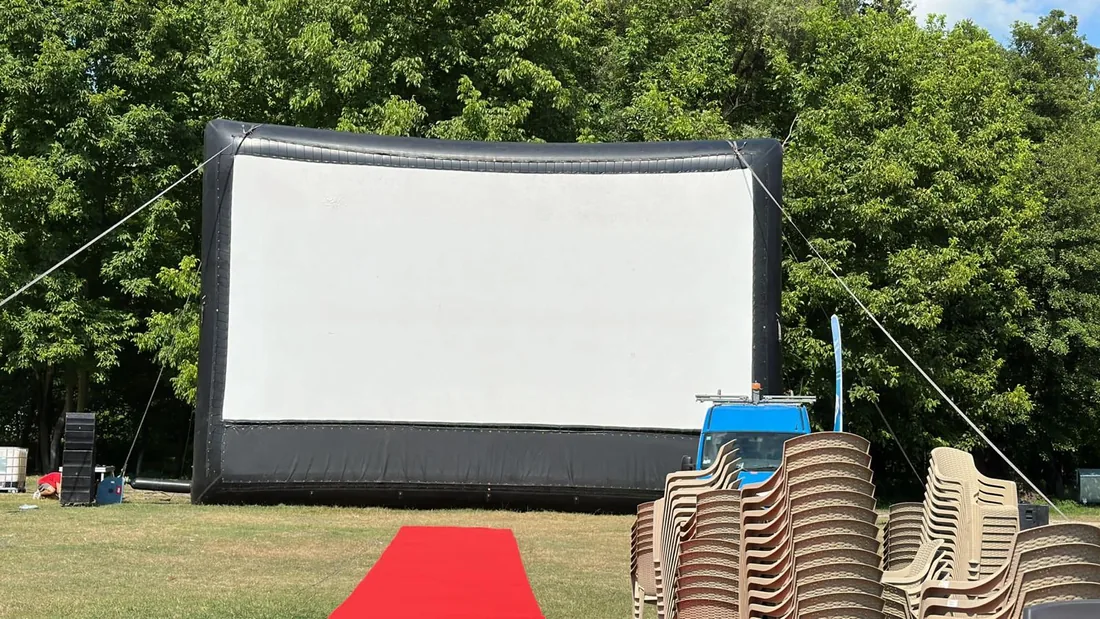 Ecran de cinema în aer liber montat printre copaci, cu covor roșu la intrare